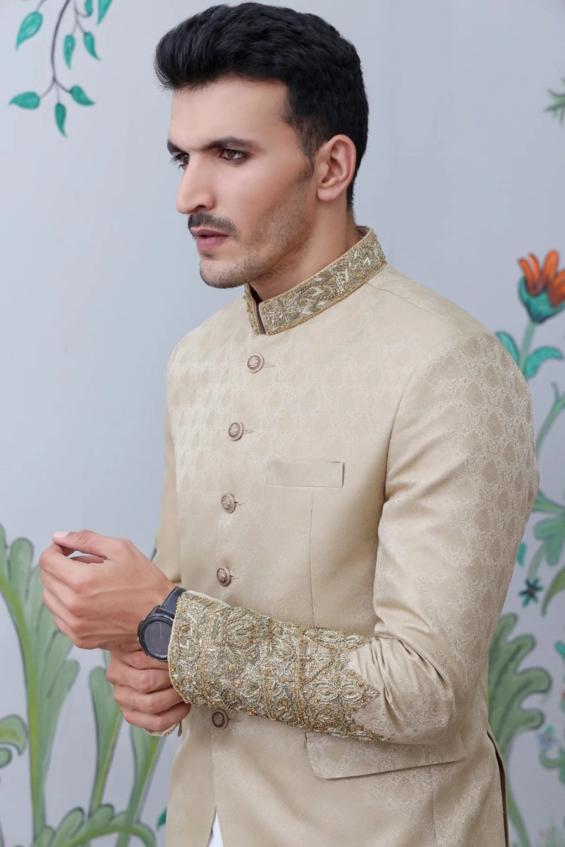 K&ouml;nnte beinhalten: Ein beigefarbener Blazer mit goldener Stickerei an Kragen und &Auml;rmeln. Der Blazer hat einen Stehkragen und einen Knopfverschluss.