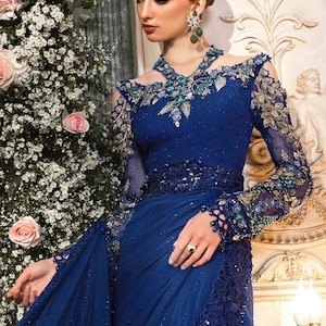 May include: A woman wearing a blue saree with intricate embroidery and embellishments. The saree has a sheer fabric with a lace border and is paired with a matching blouse. The woman is wearing a necklace and earrings with green gemstones.