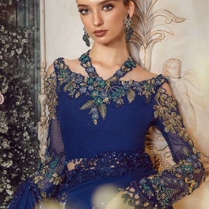 May include: A woman wearing a blue and gold embroidered saree with a matching blouse. She is wearing a necklace and earrings with green and blue stones.