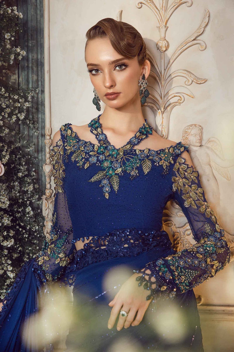 May include: A woman wearing a blue and gold embroidered saree with a matching blouse. She is wearing a necklace and earrings with green and blue stones.