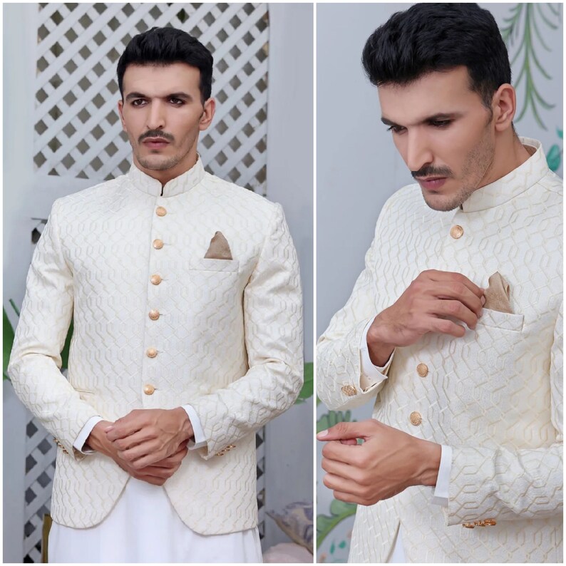 May include: Cream-coloured formal jacket with a textured pattern and gold-coloured buttons. The jacket features a mandarin collar and a pocket square. The sleeves are long, and the jacket is paired with white trousers. A stylish formal wear.