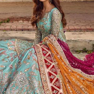 May include: A woman wearing a teal blue and gold embroidered dress with a long, flowing dupatta in shades of orange, red, and pink. The dupatta has intricate gold embroidery and a velvet border.