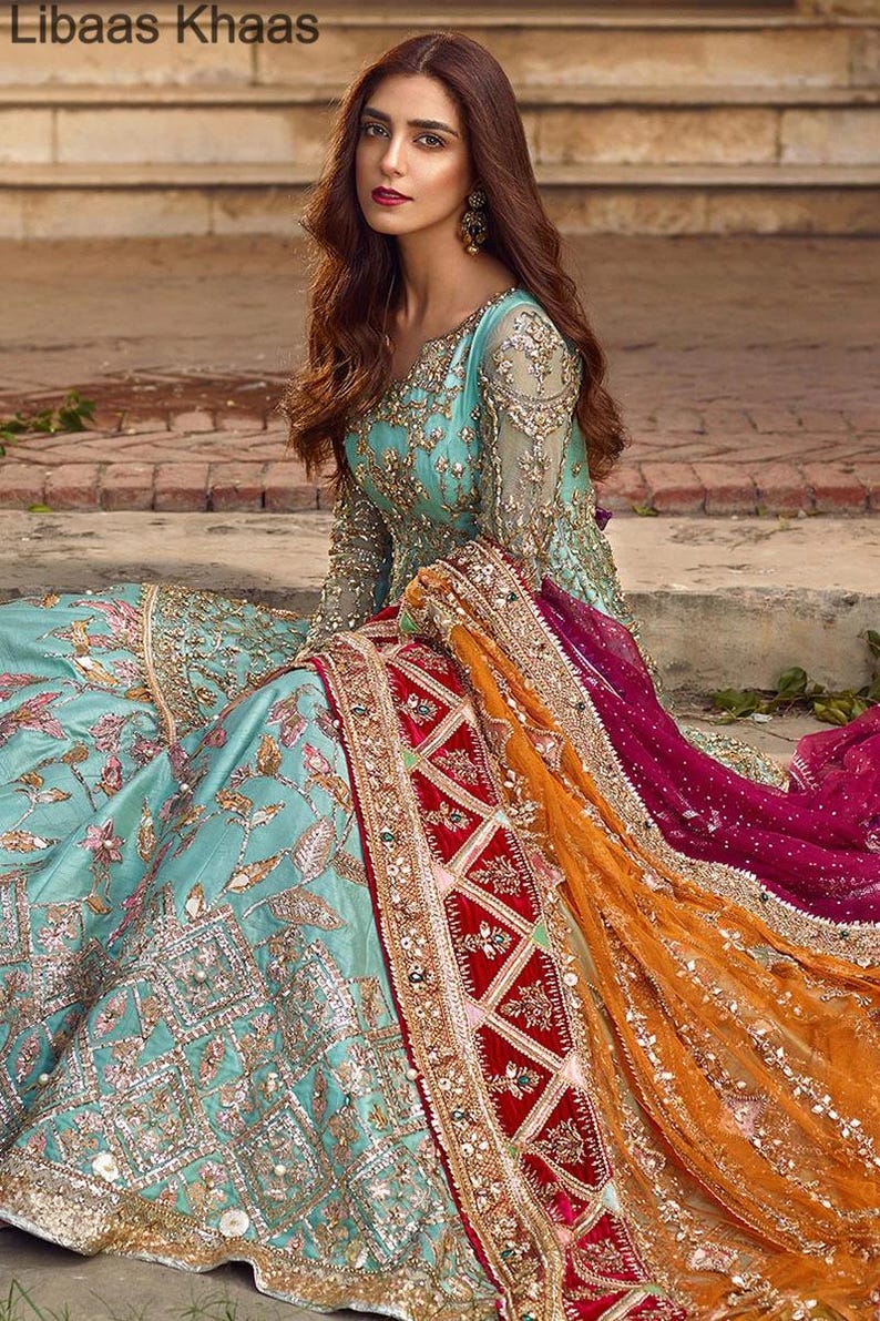 May include: A woman wearing a teal blue and gold embroidered dress with a long, flowing dupatta in shades of orange, red, and pink. The dupatta has intricate gold embroidery and a velvet border.