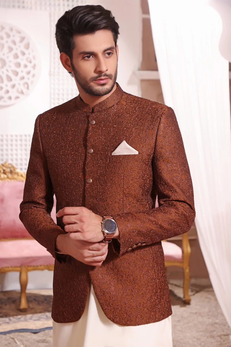 May include: A brown patterned blazer with a mandarin collar and a single button fastening. The blazer has a pocket on the left side with a white handkerchief. The blazer is worn over a white shirt and trousers.