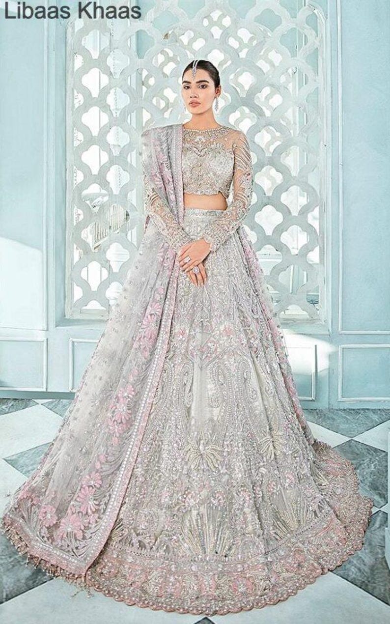 May include: A silver and pink bridal lehenga with intricate embroidery and embellishments. The lehenga has a long, flowing skirt and a fitted bodice. The dupatta is a sheer fabric with a delicate floral pattern.