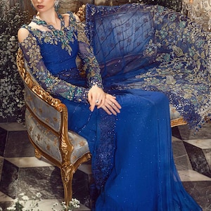 May include: A woman wearing a royal blue gown with intricate gold and green embroidery. The gown has a sheer overlay and a plunging neckline. She is sitting on a gold and blue ornate chair in a room with a vintage aesthetic.