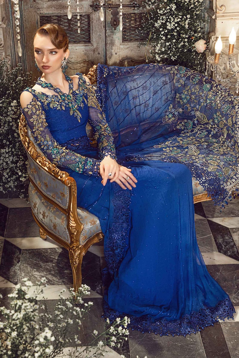 May include: A woman wearing a royal blue gown with intricate gold and green embroidery. The gown has a sheer overlay and a plunging neckline. She is sitting on a gold and blue ornate chair in a room with a vintage aesthetic.