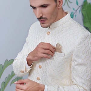 May include: Cream-coloured jacket with a geometric pattern, mandarin collar, and gold buttons. A pocket square adds a touch of elegance. The jacket is part of a formal wear ensemble.