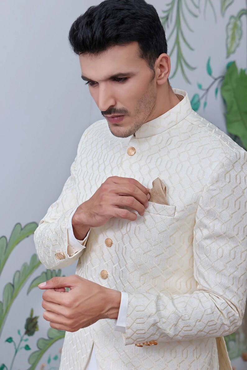 May include: Cream-coloured jacket with a geometric pattern, mandarin collar, and gold buttons. A pocket square adds a touch of elegance. The jacket is part of a formal wear ensemble.