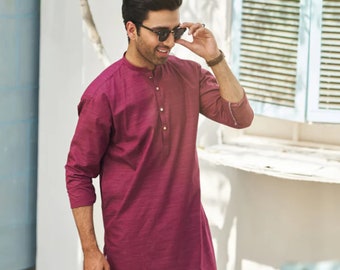Op de afbeelding: Een man draagt een lang mouw shirt in bordeauxrood met een knoopsluiting en een witte broek. Hij draagt een zonnebril en glimlacht.