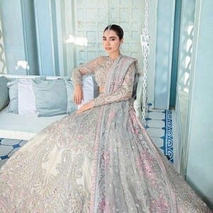 May include: A woman wearing a silver and pink embroidered dress with a long train. The dress has a sheer overlay with floral patterns. The woman is sitting on a white couch with a blue background.