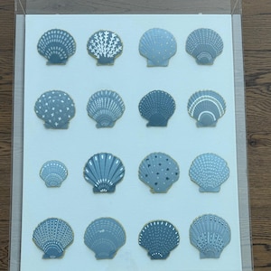 May include: A rectangular clear acrylic display case containing sixteen individual scallop shells arranged in a four-by-four grid. Each shell is painted in various shades of blue and white with unique patterns, including dots, lines, and stars.