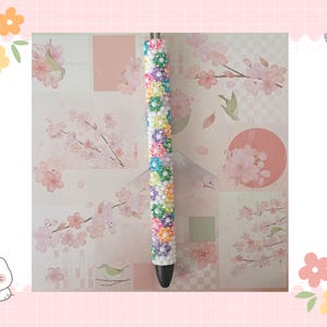Könnte beinhalten: Ein zylindrischer Stift mit einem Schaft, der mit kleinen, bunten blumenförmigen Perlen in Rosa, Blau, Gelb und Lila auf weißem Grund bedeckt ist. Der Stift hat eine schwarze konische Spitze und einen silberfarbenen Mechanismus am oberen Ende.