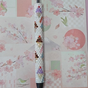 Puede incluir: Un bolígrafo blanco decorado con pequeñas cuentas blancas y pedrería de colores. El bolígrafo presenta varios diseños de conos de helado con acentos morados, marrones y rojos. El fondo es un estampado floral rosa.