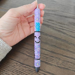 May include: A decorative pen covered in iridescent purple rhinestones, with a section of blue rhinestones and black accents. The pen has a silver tip and a black ink cartridge.