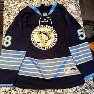 May include: A dark navy blue Reebok hockey jersey featuring a light blue collar, white laces, and the Pittsburgh Penguins logo in the center. The sleeves display the number 8 on the right and 5 on the left, with light blue stripes and a size L/XL tag.
