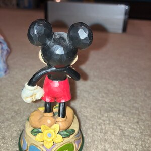 Pu&ograve; includere: Una statuetta in ceramica dipinta a mano di Topolino con pantaloncini rossi e scarpe nere. &Egrave; in piedi su una base colorata con un fiore giallo.
