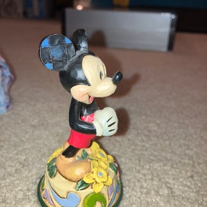 Pu&ograve; includere: Una statuetta in ceramica di Topolino in piedi su una base colorata con fiori gialli. Topolino indossa pantaloncini rossi e un guanto bianco. Il suo orecchio &egrave; a scacchi blu e neri.