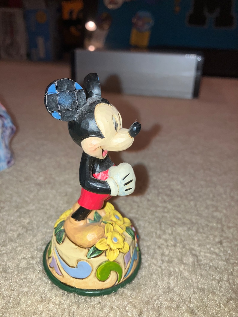 Pu&ograve; includere: Una statuetta in ceramica di Topolino in piedi su una base colorata con fiori gialli. Topolino indossa pantaloncini rossi e un guanto bianco. Il suo orecchio &egrave; a scacchi blu e neri.