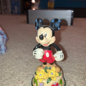 Pu&ograve; includere: Una statuetta in ceramica dipinta a mano di Topolino in piedi su una base colorata con fiori. Topolino indossa i suoi pantaloncini rossi caratteristici e guanti bianchi.