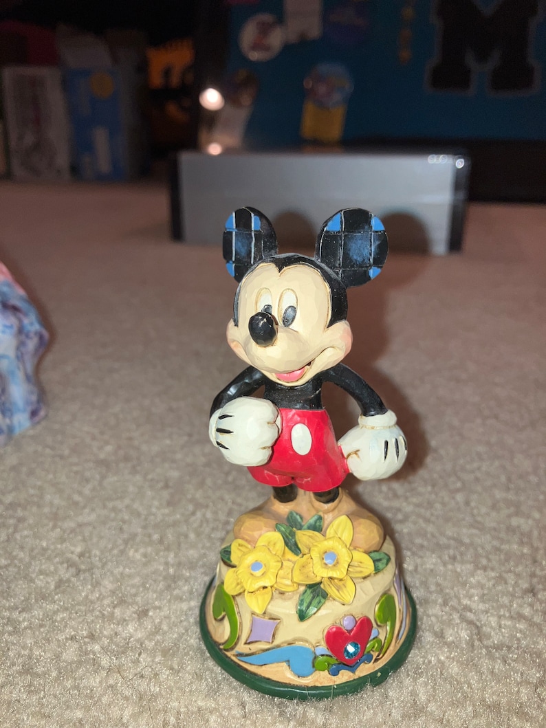Pu&ograve; includere: Una statuetta in ceramica dipinta a mano di Topolino in piedi su una base colorata con fiori. Topolino indossa i suoi pantaloncini rossi caratteristici e guanti bianchi.