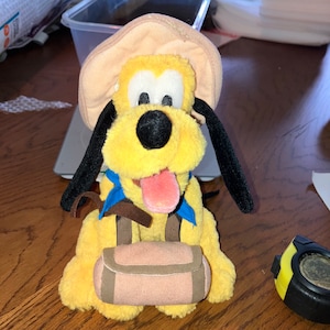 May include: A yellow plush toy dog wearing a brown hat and a blue bandana. The dog is carrying a brown backpack.