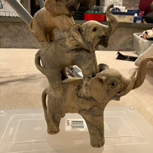 May include: A stacked elephant figurine. Three elephants are balanced on top of each other, with their trunks raised. The elephants are a light beige color with darker gray marbling.