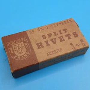 Puede incluir: Una caja rectangular de cartón de remaches partidos, con el texto "SPLIT RIVETS" en un lugar destacado. La caja es marrón y beige con el logotipo "Buckeye R&S Co. BRAND" en el lado izquierdo. Contiene 50 remaches cobrizados No. 1.