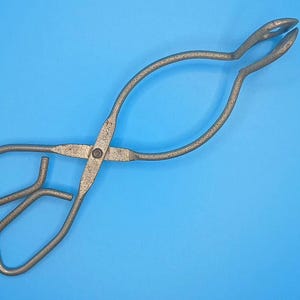 Può includere: Pinze in metallo vintage con design a forbice, manico curvo ed estremità di presa. Le pinze sono in metallo grigio usurato e sono poste su uno sfondo blu uniforme. Lo strumento viene probabilmente utilizzato per la manipolazione di oggetti caldi.