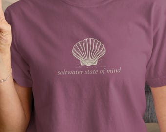 May include: A mauve short-sleeved t-shirt featuring a white line-art illustration of a seashell centered on the chest. Below the seashell, the text "saltwater state of mind" is printed in a simple, lowercase white font on the front of the garment.