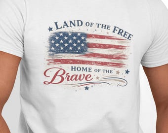 May include: A white short-sleeved t-shirt featuring a distressed American flag graphic in the center. Above the flag, the text reads LAND OF THE FREE in blue, and below the flag, the text reads HOME OF THE Brave in red and blue script.