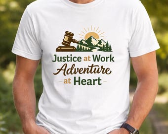 May include: White t-shirt featuring a graphic with a gavel, mountains, and sun. The text reads "Justice at Work Adventure at Heart" in brown and green. The design evokes themes of law and outdoor adventure.