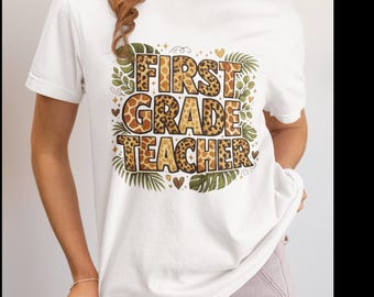May include: A white short-sleeved t-shirt featuring the text FIRST GRADE TEACHER in a bold, blocky font with a brown and tan leopard print pattern. The design is surrounded by green tropical palm leaves, small hearts, and decorative star accents.