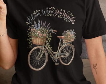 May include: A black t-shirt featuring a graphic of a bicycle with a front wicker basket and a rear wooden crate, both filled with colorful flowers. The text "Bloom Where You Wander" is printed in a cursive font above the bicycle illustration.