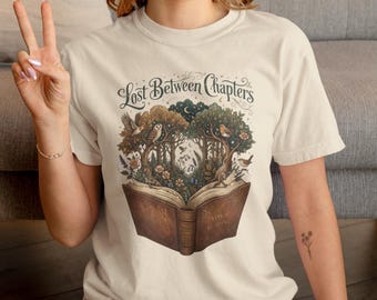 May include: A cream-colored short-sleeve t-shirt featuring a graphic of an open book with a forest growing out of its pages. The text "Lost Between Chapters" is printed above the illustration, and "Magic Reading" is on the book cover.