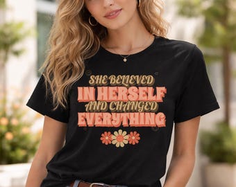 May include: Black t-shirt with the text "SHE BELIEVED IN HERSELF AND CHANGED EVERYTHING" in a retro font. The design includes flower graphics in shades of orange and yellow. The shirt has short sleeves and a crew neck.