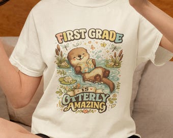 May include: A cream-colored t-shirt featuring a graphic of an otter floating in water while reading a book. The text reads FIRST GRADE IS OTTERLY AMAZING. The design includes cattails, water lilies, dragonflies, and small butterflies in soft colors.