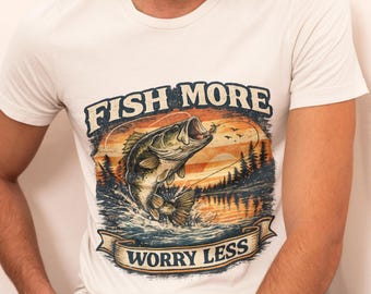 May include: A cream-colored short-sleeve t-shirt featuring a graphic print of a jumping bass fish over a lake at sunset. The text FISH MORE is arched above the fish, and WORRY LESS is printed on a banner below, both in dark blue lettering.