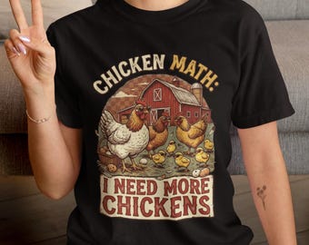 May include: A black short-sleeved t-shirt featuring a graphic of a red barn, several chickens, and chicks. The text reads "CHICKEN MATH: I NEED MORE CHICKENS" in a vintage-style font. The design includes a basket of eggs and scattered eggs.