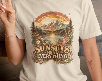 May include: A cream-colored short-sleeve t-shirt featuring a graphic print of a mountain landscape at sunset with pine trees, flowers, and birds. The text "SUNSETS HEAL EVERYTHING" is printed in a bold, stylized font at the bottom of the design.