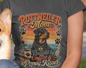 May include: A dark gray t-shirt featuring a graphic print of a Rottweiler dog, a coffee mug, and paw prints. The text reads "ROTTWEILER Mom" and "Powered by Coffee & Puppy Kisses" in a retro-style font over a multi-colored sunset background.