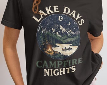 May include: A black t-shirt featuring a circular graphic of a mountain landscape at night with a campfire, a lake, and a canoe. The text "LAKE DAYS & CAMPFIRE NIGHTS" is printed in a cream-colored font above and below the circular illustration.