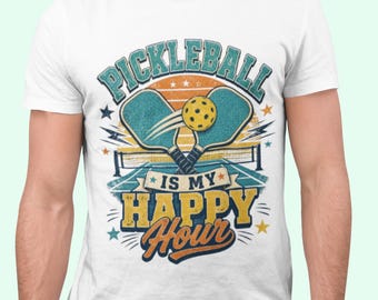 May include: A white short-sleeve t-shirt featuring a graphic print of two teal pickleball paddles and a yellow ball over a court. The text reads PICKLEBALL IS MY HAPPY HOUR in blue and yellow lettering with a sunset background and small stars.