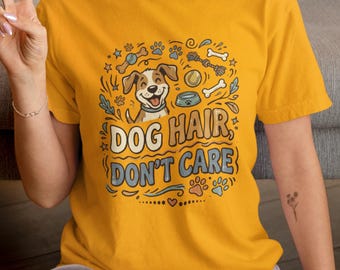 May include: A mustard yellow t-shirt featuring a graphic illustration of a smiling dog surrounded by bones, a ball, and a food bowl. The text "DOG HAIR, DON'T CARE" is printed in the center in a bold, stylized font, accented by paw prints and swirls.