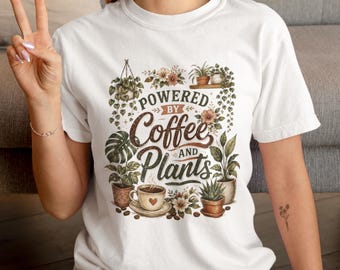 May include: A white short-sleeve t-shirt featuring a graphic print of various potted plants, a coffee cup, and the text "POWERED BY Coffee AND Plants" in brown and green tones. The design includes hanging plants, a monstera leaf, and coffee beans.