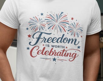 May include: A white short-sleeved t-shirt featuring a graphic print of red and blue fireworks. The text reads Freedom IS WORTH Celebrating in a mix of blue and red script and block lettering, accented by small red and blue stars.