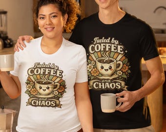 May include: A white short-sleeved t-shirt and a black short-sleeved t-shirt, both featuring a graphic print of a smiling coffee cup surrounded by flowers and sunburst rays, with the text "Fueled by COFFEE and CHAOS" printed in a vintage style.