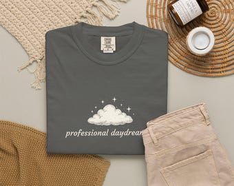 May include: A dark gray t-shirt featuring a white graphic of a cloud surrounded by small four-pointed stars. Below the cloud, the text reads "professional daydreamer" in a white, lowercase, sans-serif font. The shirt has a Comfort Colors label.