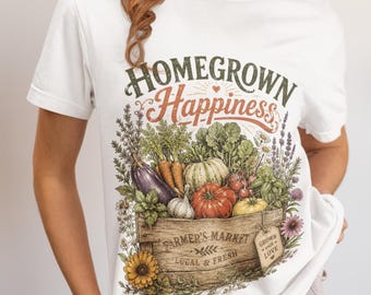 May include: A white t-shirt featuring a graphic of a wooden crate filled with fresh vegetables including carrots, tomatoes, and an eggplant. The text reads "HOMEGROWN Happiness" at the top and "FARMER'S MARKET LOCAL & FRESH" on the crate.