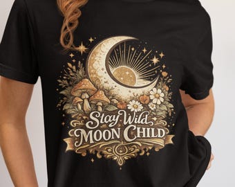 May include: A black short-sleeved t-shirt featuring a graphic print of a crescent moon, sun rays, mushrooms, and flowers. The text "Stay Wild MOON CHILD" is printed in a decorative font over a banner design in shades of gold, beige, and brown.